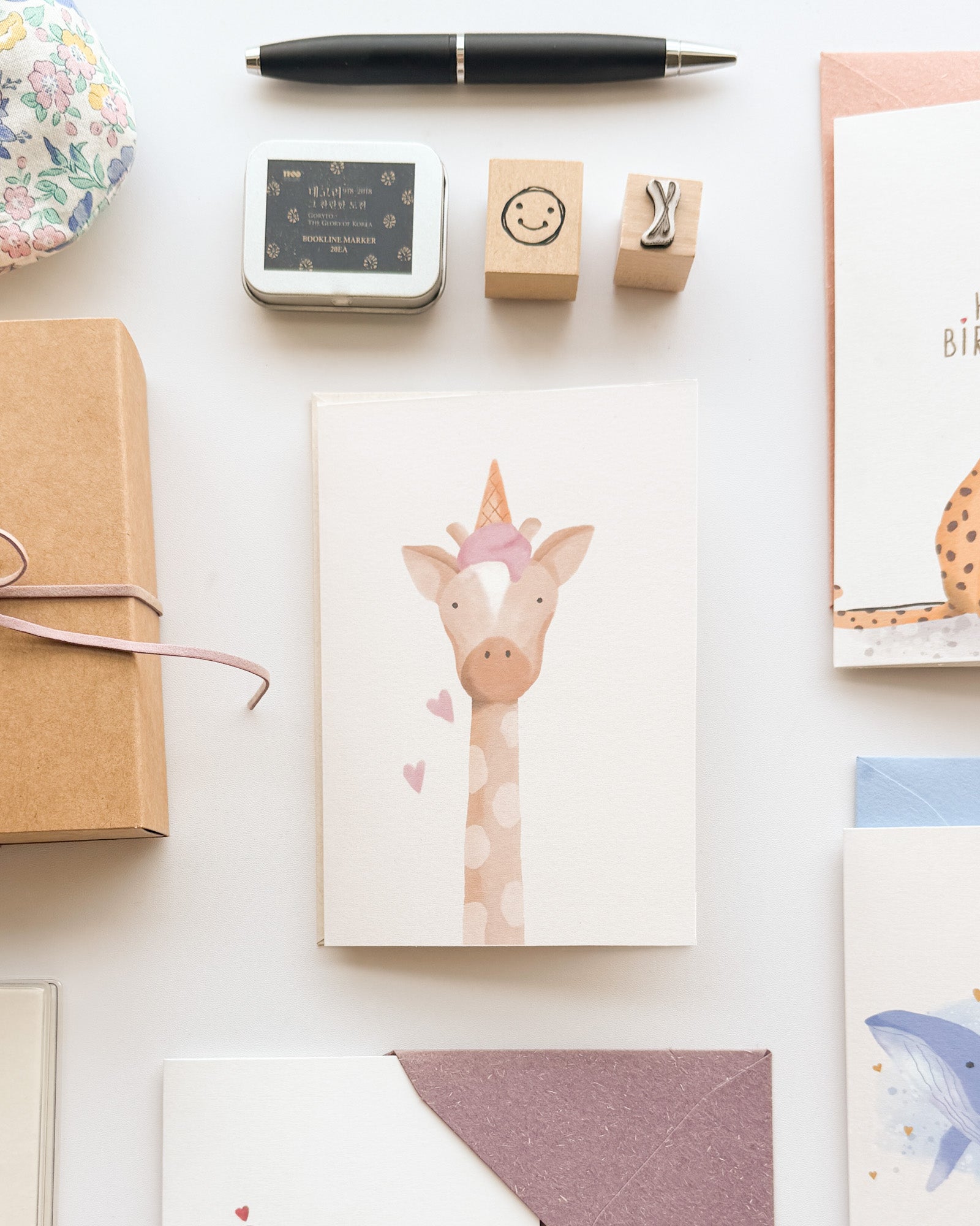 Stationery items including a giraffe card, gift box, and pen on a white surface
