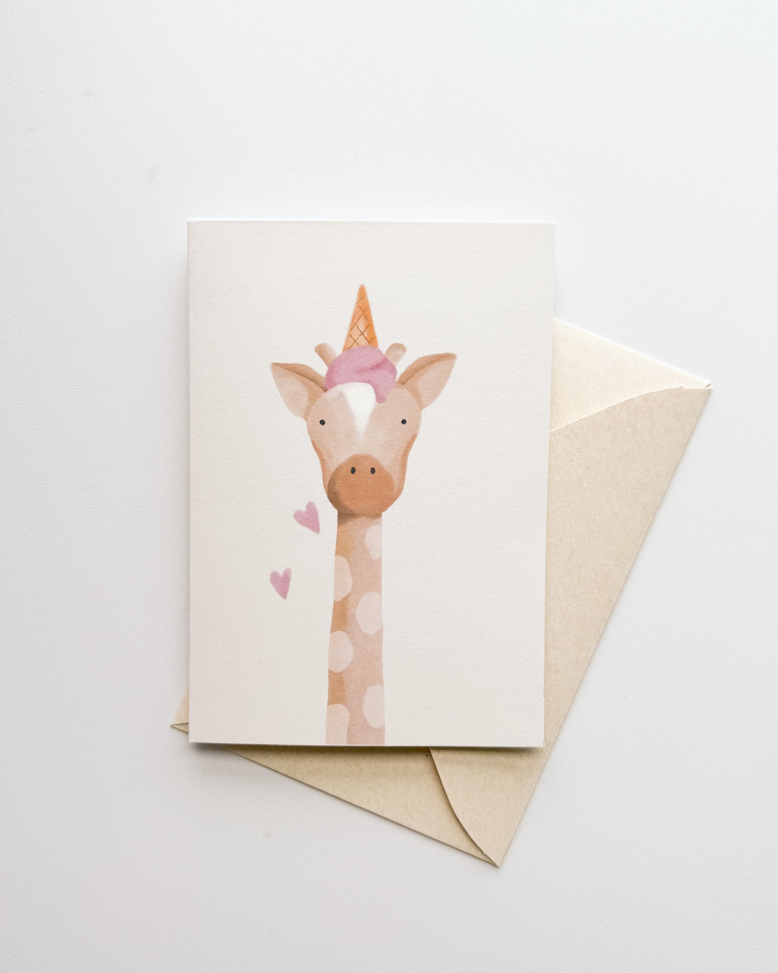 Greeting card with a giraffe illustration with an ice-cream on top of its head, accompanied by a beige envelope on a light gray background.